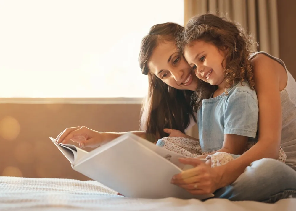 mom and daughter smiling and bonding while reading a book in bed